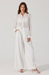 A woman is wearing a sheer, long-sleeved white blouse with a subtle floral pattern and a classic shirt collar. The blouse is buttoned down the front, with the top few buttons left open. She pairs the blouse with high-waisted, wide-leg white trousers that have a tailored fit and pleats at the waist. She completes her outfit with white high-heeled sandals featuring a thin strap across the toes and an ankle strap.